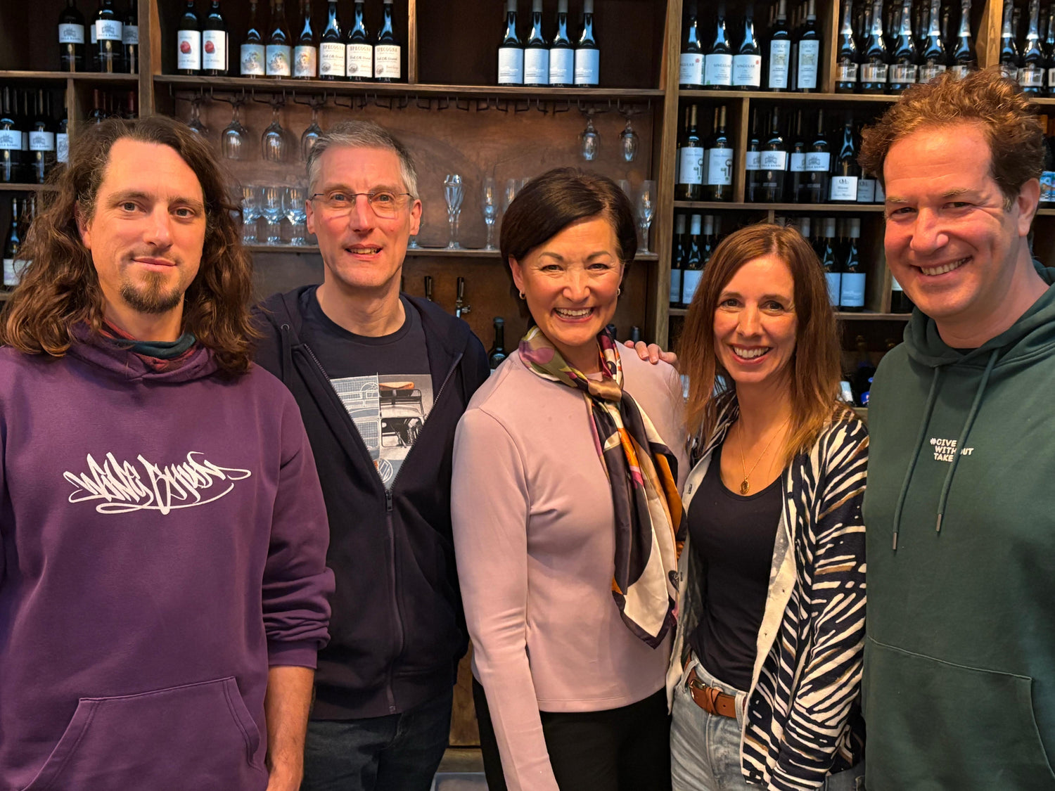 Group of five people posing together in a wine cellar with shelves of wine bottles in the background.